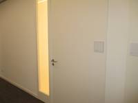 weiße  Tür in einer weißen Wand, links neben der Tür ist eine schmale Glasscheibe, rechts etwa in Kopfhöhe ein Hinweisschild