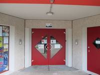 Eingang mit roter zweiflügeliger Drehflügeltür, in der Mitte der Türen sind dreieckige Glasfenster