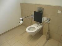 Eine weiße Behindertentoilette an einer gekachelten Wand. Haltegriffe rechts und links von der Toilette.
