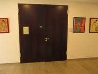 braune geschlossen Tür in einer hellen Wand. Auf der linken Tür ist ein Aushang, rechts und links von der Tür hängen verschiedene Bilder