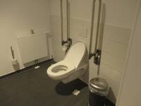 Eine weiße Toilette an einer weiß gefliesten Wand. Der Boden ist dunkel. Bei der Toilette ist auf jeder Seite ein Haltegriff.