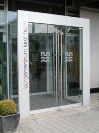 zweiflüglige gläserne Eingangstür mit dem Logo der Stadt Heidelberg darauf, die Türen sind umgeben von einem metallischen Rahmen mit dem Schriftzug Bürgerzentrum Kirchheim, der Eingang ist etwas hervorgesetzt vom Gebäude