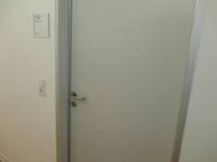 geschlossene hellgraue Tür in einer weißen Wand. Links von der Tür ein Lichtschalter und ein Türschild.