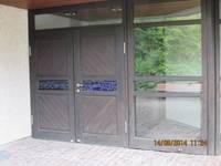 Zweiflügelige hölzerne Eingangstür, links davon eine große Glasscheibe, der Platz davor ist aus Kleinsteinpflaster 
