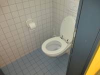  weiße Toilette an einer weiß gekachelten Wand, links an der Wand hängt eine WC-Papierrolle.