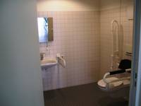 Behindertentoilette mit grauen Boden und weiß gekachelten Wänden, rchts ist der Toilettensitz mit zwei Haltegriffe und links das Waschbecken zu sehen