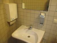 Waschbecken an einer gekachelten Wand, darüber ein Spiegel, links ein Papierhandtuchhalter, rechts an der Wand ein Seifenspender