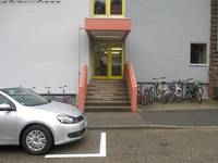 gelb gerahmte Glastür mit Überdachung, davor Stufen mit Handlauf beidseitig, Fahrräder rechts und links, im  Vordergrund Straße