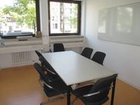 Ein Raum mit einem großen Tisch und 6 Stühlen darum. An der Rückseite sind zwei Fenster mit Heizkörper darunter. An der rechten Wand ist ein Whiteboard. Der Boden ist orange.