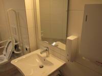 Ein weißes Waschbecken an einer weiß gekachelten Wand, darüber ein Spiegel, links neben dem Waschbecken ist die Behindertentoilette.