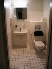 weiß gekachelte Behindertentoilette, an der hinteren Wand Toilettensitz, Waschbecken und Papierhandtuchspender, keine Haltgriffe