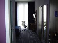 Auf dem Bild sieht man einen Ausschnitt des Raums vom Eingang aus gesehen, links vorne ist die Tür zum Bad rechts ein Spiegel und eine Tür zu einem2. Zimmer, weiter hinten im Bild ist ein Stuhl, ein Tisch und ein Stück vom Bett zu erkennen, über dem Tisch befindet sich ein Fernseher