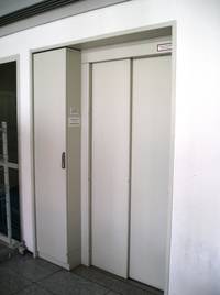 der weiße geschlossene Aufzug ist zu sehen, links ist der Ruftaster, um den Aufzug ist etwas von der Gebäudewand und von dem Boden zu erkennen