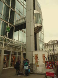 Gläserner Außenaufzug in Fahrt zwischen den Stockwerken, unten ein Teil des Bismarckplatz, weiter oben Zugang zum Gebäude über einen überdachten Steg 