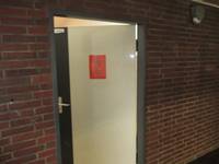 geöffnete weiße Tür in einer Backsteinwand, auf der Tür ein rotes Schild