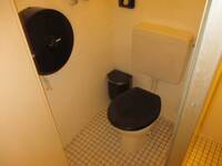 Toilette mit scharzem Deckel in einem weiß gekacheltem Raum. Links neben der Toilette steht ein schwarzer Mülleimer, an der Wand links hängt ein großer, runder WC-Papierspender asu schwarzem Kunststoff
