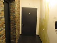  dunkle Tür in einer hellen Wand, davor Flur. In der linken Wand ist eine geschlossene Aufzugstür
