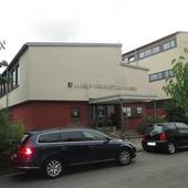 zwei beieinander stehende Gebäude, auf dem linken Gebäude Schriftzug "Albert Schweitzer Schule" und ein überdachter Eingang mit Stufen, davor Straße mit parkenden Autos