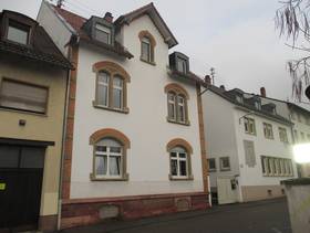 zweistöckiges, weißes Mehrfamilienhaus mit durch Bögen verzierten Fenster, rechts und links weitere Gebäude, davor eine Straße
