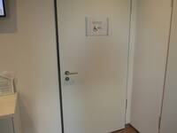 Eine helle Tür mit einem dunklem Rahmen in einer weißen Wand. Auf der Tür befindet sich ein Schild mit der Aufschrift "WC" und einem Rollstuhlsymbol.