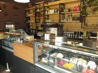 Eine Holztheke zwischen zwei niedrigen Glasvitrinen mit Speiseeis und Backwaren. Im Hintergrund sind offene Regale mit Geschirr, Pappbechern, Deko, Kaffee und Pflanzen