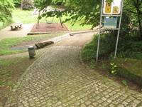 gepflasterter Weg mit leidter Rechtskurve. Rechts davon Bäume, Büsche und ein Schild. Im Hintergrund eine Schaukel und eine steinerne Tischtennisplatte.