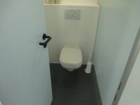 weißes Hänge-WC an einer weißen Wand und einem dunklen Boden. Über der Toilette ist der Spültaster, rechts ist eine weiße Trennwand und eine WC-Bürste auf dem Boden. Links die WC-Tür, die nach innen geöffnet ist