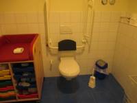 weißes Hänge-WC an weiß gekachelten Wand mit Haltegriffen rechts und links, Spülung an der Wand hinter WC-Sitz. Auf dem blauen Fliesenboden rechts von  Toilette  Mülleimer und WC-Bürste, links steht ein Wickeltisch