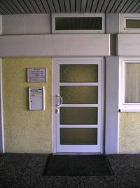 weiße mit Glaselementen versehene Tür an einer gelblichen Wand, links Praxisschild und Briefkasten, rechts Fensterausschnitt, im Vordergrund Natursteinplatten und Bodenmatte