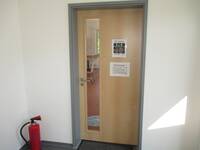 Tür in heller Holzoptik, im linken DRittel langes schmales Glaselement.
Links in der Ecke vor der Tür steht auf dem Boden ein roter Feuerlöscher