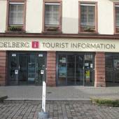 mehrstöckiges Gebäude mit einer Ladefront mit zwei Glasschiebtüren. Darüber Schriftzug "Heidelberg Tourist Information". Vor der Tür großes Podest als Abgrenzung zum Platz