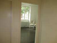 Eine offenstehende Tür in einem weißen Rahmen, in einer weißen Wand, dahinter kleiner Raum mit dunklem Bodenbelag, Fenster und 2 Stühlen