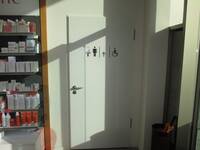 Eine weiße Türe in einer weißen Wand. Links neben der Tür sind Verkaufsregale mit Waren. Auf der Tür sind Symbole: Mann, Frau, Rollstuhlnutzende und Wickeltisch