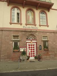 Ausschnitt mit rotgepunkteten Eingangstür, auf beiden Seiten Fenster, vor dem Eingang eine Stufe und Pflanzenkübel, über dem Eingang im 1. Obergeschoss das Bild einer Frau