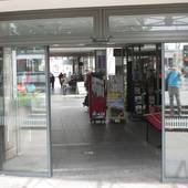 gläserner Eingangsbereich mit automatischer Schiebetür, dahinter ein Flur mit Steinfliesen, rechts ein Laden