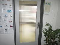 einflügelige Glastür mit weißem Rahmen und dunkler Zarge. Rechts neben der Tür ist ein DIN-A4 Schild mit den Öffnungszeiten. Links von der Tür sind mehrere Aushänge