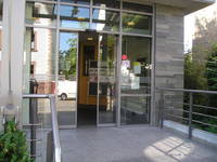 Glasfront mit automatischer geöffneter Schiebetür, rechts grauer Gebäudeteil, im Vordergrund Bewegungsfläche vor Eingang mit Geländer