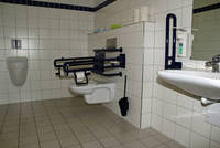 Behindertentoilette weiß gefliest, rechts erst Waschbecken, danach Toilette mit Haltegriffen