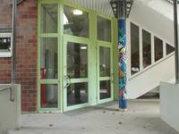 eine gläserne, einflügelige Eingangstür in einem grünen Rahmen, rechts daneben eine weitere ähnliche Tür, links daneben und darüber weiter Glasscheiben, davor eine Bewegungsfläche und eine buntbemalte Säule
