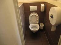 Eine weiße Toilette an einer dunklen Wand, an der rechten Wand hängt ein großer, runder WC-Paper-Spender