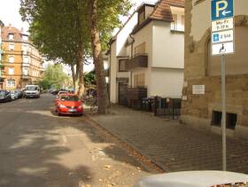 Eine Straße mit parkenden Autos. Vor einem Haus ein Behindertenparkplatz mit Schild und Bodenmarkierung