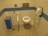 Eine weiße Toilette an einer weiß gekachelten Wand mit einem blauen Streifen rings an der Wand. Links ist ein Haltegriff.