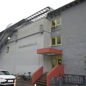 zweiteiliges Flachdachgebäude, linker Teil ohne Fenster mit einer Feurleiter und einem Schriftzug, der rechte Teil etwas zurückgesetzt mit einem überdachten Eingang, Stufen und Fenstern mit gelben Rahmen