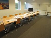 Ein großer Raum mit einer langen Tischgruppe und Stühlen, an der hinteren Wand hängt ein Whiteboard
