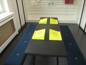 langer schwarzer Tisch, darauf stehen 4 gelbe, geneigte Auflagestationen (Sharepoints) für iPads. Links und rechts vom Tisch stehen zwei blau gepolsterte Bänke ohne Rückenlehne