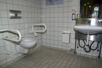 gefliester weißer Raum, links Toilette mit Haltegriffen, rechts Waschbecken