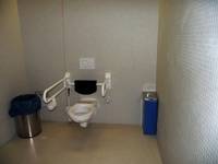 Gefliester Raum; Toilette mit Rückenlehne, Drückerplatte an Wand über Rückenlehne, Hygienebehälter an linker Umsetzfläche, Mülleimer an rechter Umsetzfläche