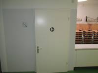 helle Tür vor heller Wand, auf Tür Symbol für Wickeltisch, neben deer Tür Schild für Behindertentoilette, rechts von Tür ein Stück der Gaderobe
