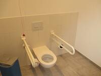 Eine weiße Toilette, mit je einem Haltegriff auf jeder Seite, in einer weiß gekachelten Wand. Der Boden hat Fliesen in beige