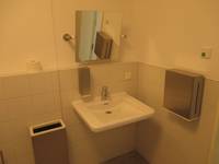 weißes Waschbecken an einer hell gekachelten Wand, rechts davon ein Papierhandtuchhalterc, über dem Becken ein Spiegel und ein Seifenspender